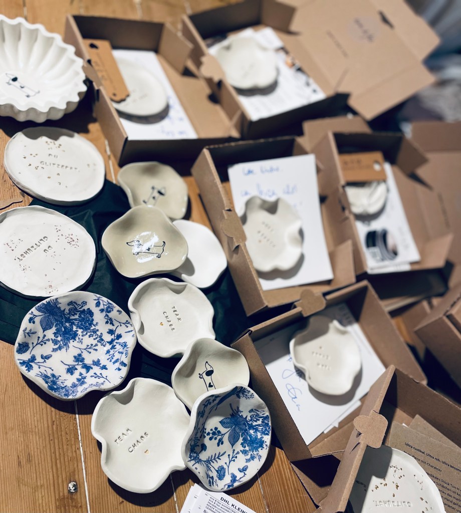 Blick ins Atelier einer Keramikmanufaktur mit echtem Verpackungschaos. Kartons, Seidenpapier, Etiketten, Bänder und fertige Keramikstücke liegen auf dem Arbeitstisch. Zwischen Verpackungsmaterial, Tonresten und Werkzeugen entstehen handgemachte Keramikprodukte für den Online-Shop. Authentischer Arbeitsalltag einer selbstständigen Keramikerin beim Verpacken und Vorbereiten von Bestellungen, echtes Handwerk hinter den Kulissen, kein perfektes Studio, sondern echtes Arbeiten mit Ton.